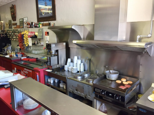 A diner kitchen with a grill, pots, and utensils, featuring a colorful wall of snacks and drinks in the background.