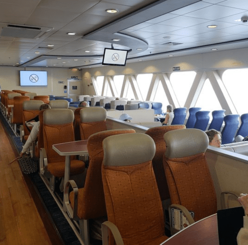 Interior of a ferry with rows of orange and blue seats, large windows, and a no smoking sign displayed on a screen.
