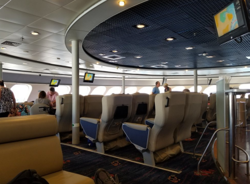 Interior of a ferry with rows of seats, large windows, and passengers mingling in a spacious, well-lit area.