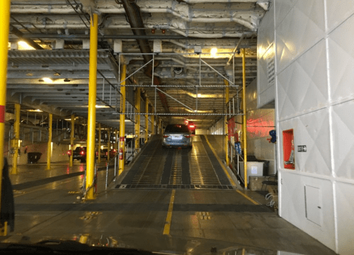 A vehicle drives up a ramp inside a large ferry, with yellow pillars and industrial surroundings visible.