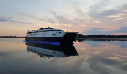 A ferry boat glides across calm waters at sunset, reflecting the colorful sky and surrounding landscape.