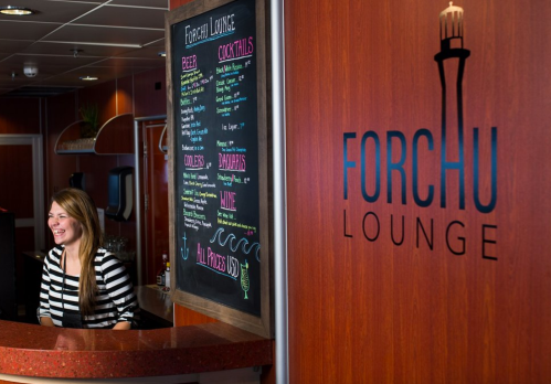 A smiling woman stands at the counter of Forchu Lounge, with a menu board displaying drinks and snacks in the background.