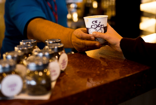 A barista hands a coffee cup to a customer at a counter, with jars of tea in the foreground.