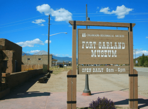 Sign for Fort Garland Museum, with hours listed as open daily from 9am to 5pm, set against a blue sky.