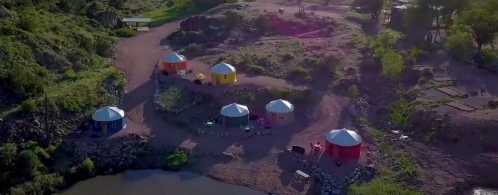 Aerial view of colorful yurts near a river, surrounded by greenery and rocky terrain.