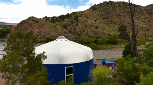 A blue yurt by a river, surrounded by green hills and trees, with outdoor seating nearby.