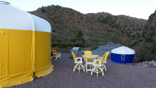 Two colorful yurts beside a river, with outdoor seating and a grill, set against a mountainous landscape.