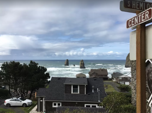 Coastal view with rocky formations, waves crashing, and houses in the foreground under a cloudy sky. Street sign visible.