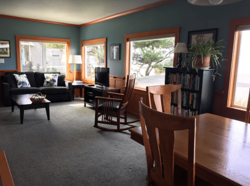 Cozy living room with a sofa, rocking chair, TV, and large windows showcasing a view of trees outside.
