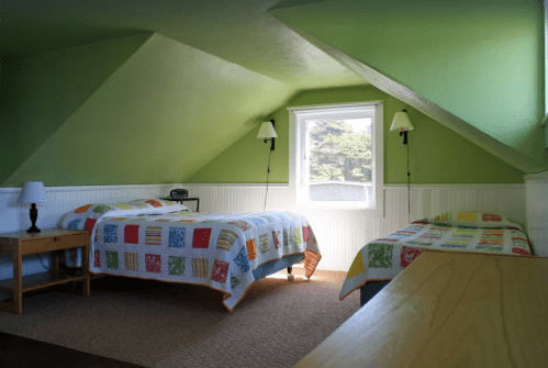 Cozy attic bedroom with two beds, green walls, colorful quilts, and a window letting in natural light.