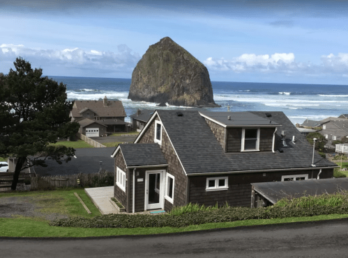 A coastal view featuring a house in the foreground and a large rock formation in the ocean under a blue sky.