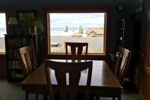 A cozy dining area with a wooden table and chairs, overlooking a beach with rock formations through a large window.