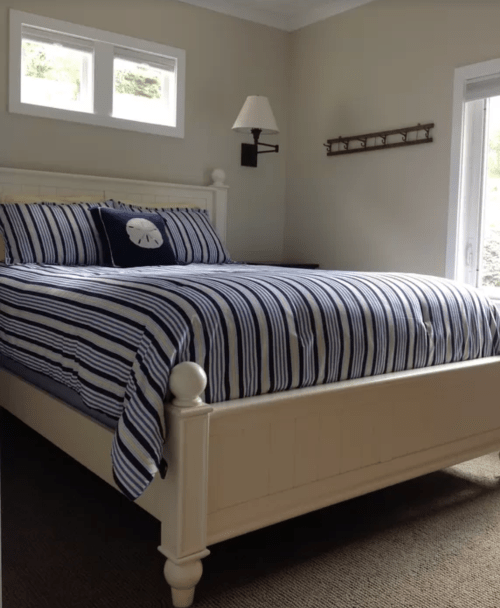 A cozy bedroom featuring a white bed with blue striped bedding and a decorative pillow, with natural light from windows.