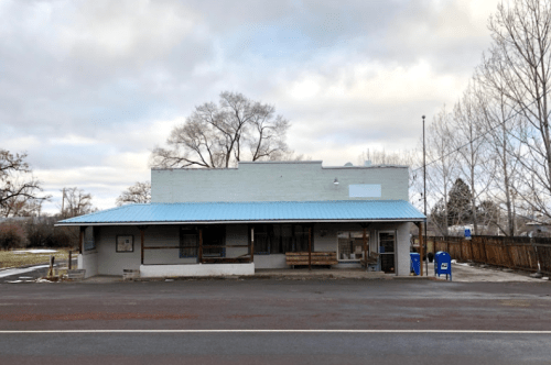 A small, single-story building with a blue metal roof, surrounded by trees and a gravel parking area.