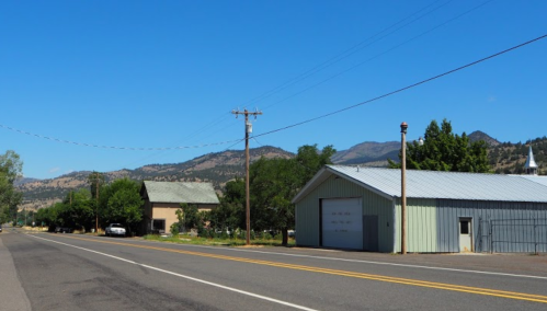A quiet rural road lined with buildings and trees, with mountains in the background under a clear blue sky.