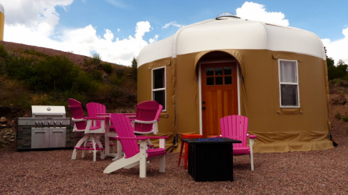 A tan yurt with a wooden door, surrounded by pink outdoor chairs and a grill, set against a blue sky with clouds.