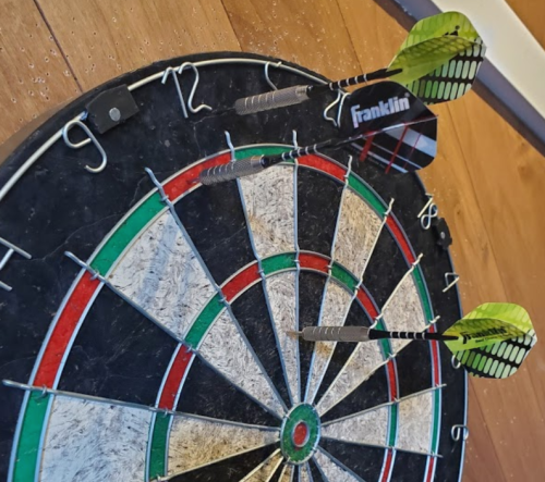 A dartboard with three darts stuck in it, featuring green and black flights, on a wooden floor.