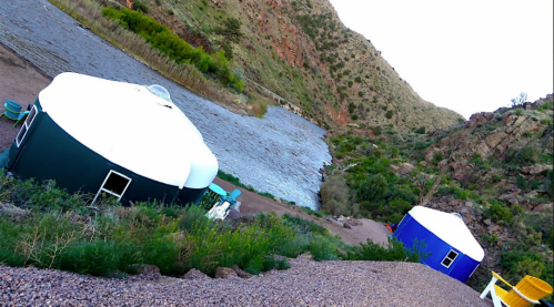 Two colorful yurts sit beside a rocky riverbank, surrounded by lush greenery and mountains in the background.