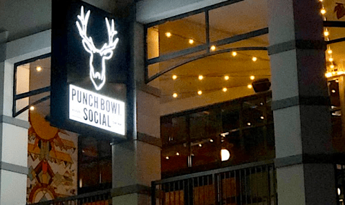A brightly lit sign for "Punch Bowl Social" featuring a deer head logo, with string lights in the background.