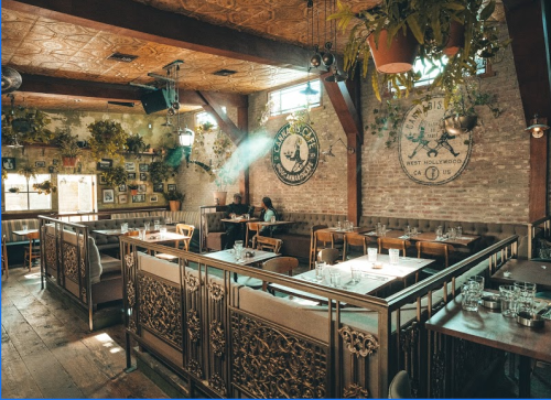 A cozy restaurant interior with wooden decor, plants, and empty tables, featuring warm lighting and a relaxed atmosphere.