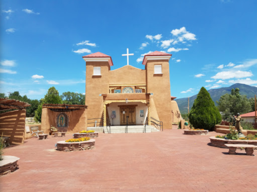 A southwestern-style church with a cross, surrounded by greenery and blue skies, featuring a brick pathway in front.