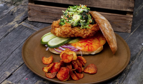 A crispy chicken sandwich topped with slaw, pickles, and tomatoes, served with a side of potato chips on a brown plate.