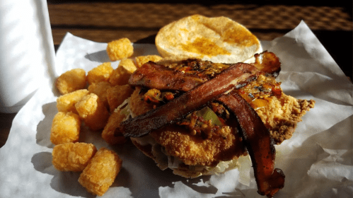 A crispy chicken sandwich topped with bacon and peppers, served with golden tater tots on the side.