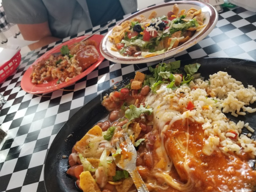 A plate of rice and beans topped with cheese and sauce, alongside a colorful salad and another dish in the background.
