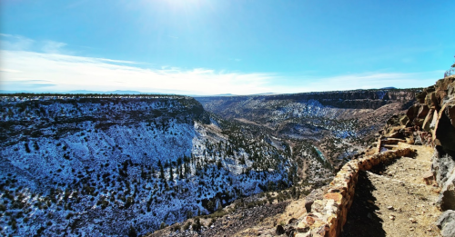 A panoramic view of a snow-covered canyon under a clear blue sky, with rocky cliffs and scattered trees.