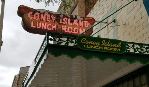 Signage for "Coney Island Lunch Room" with a neon sign above and a green sign below, set against a cloudy sky.