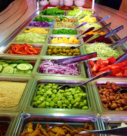A vibrant salad bar featuring a variety of fresh vegetables, fruits, and toppings in metal trays with serving utensils.