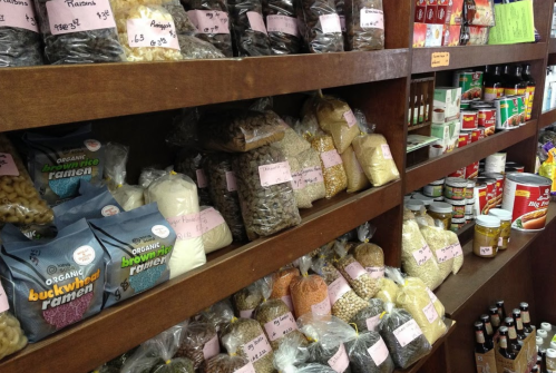 A wooden shelf filled with various packaged grains, pasta, and canned goods, all labeled with prices.