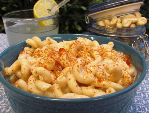 A bowl of creamy macaroni topped with paprika, a glass of lemonade with a lemon slice, and a jar of cashews in the background.