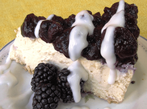 A slice of cheesecake topped with blackberry compote and drizzled with white icing, served with fresh blackberries.