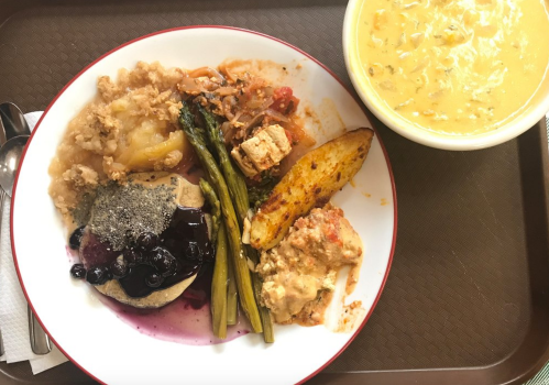 A plate with various foods: pancakes with blueberries, asparagus, potatoes, and a side of soup in a bowl.