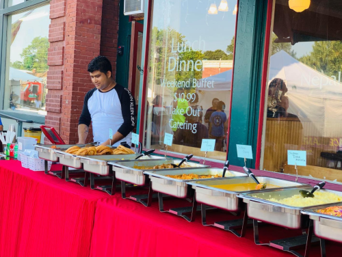 A man stands behind a buffet table filled with various dishes, with a sign for lunch and dinner options in the background.