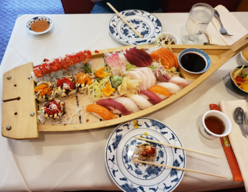 A wooden boat filled with assorted sushi, sashimi, and garnishes, accompanied by soy sauce and chopsticks on a table.