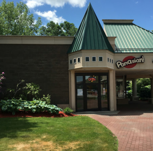 Exterior of a building with a green peaked roof, sign reading "Panasian!" and an open door, surrounded by greenery.