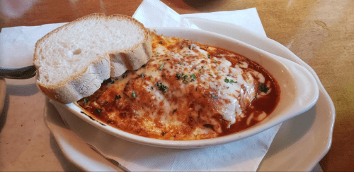 A baked dish of cheesy lasagna topped with herbs, served with a slice of bread on the side.