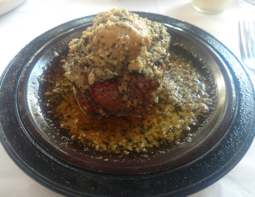 A savory meat dish topped with herbs and spices, served in a dark, round plate with a rich sauce.