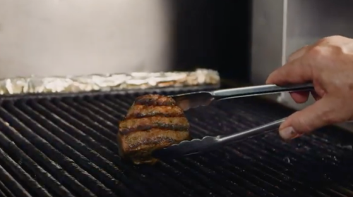A hand using tongs to flip a grilled steak on a barbecue grill with visible grill marks.