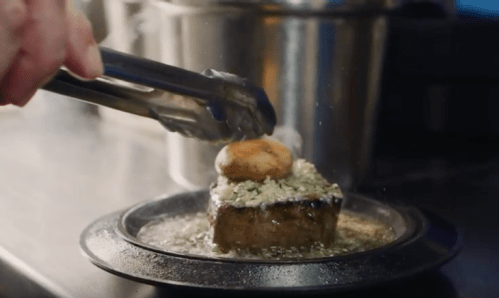 A chef uses tongs to place a biscuit on top of a steak garnished with herbs, sizzling in a pan.