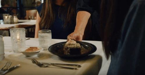 A hand serves a plate of food at a restaurant table, with drinks and dessert nearby.