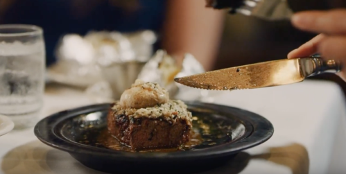 A slice of cake topped with ice cream and nuts, served on a dark plate with a fork and knife nearby.