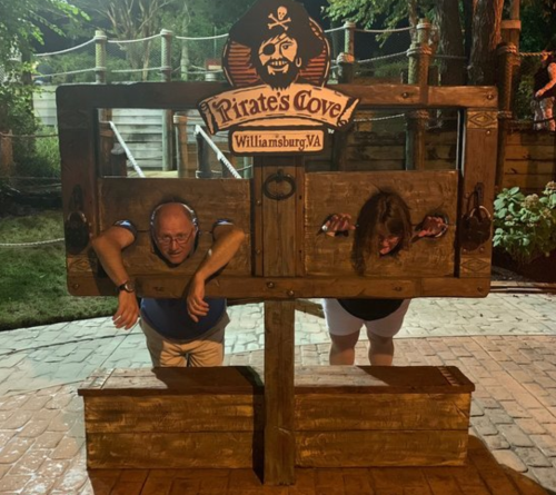 Two people are playfully posing in a pirate-themed stocks at Pirate's Cove in Williamsburg, VA, smiling for the camera.