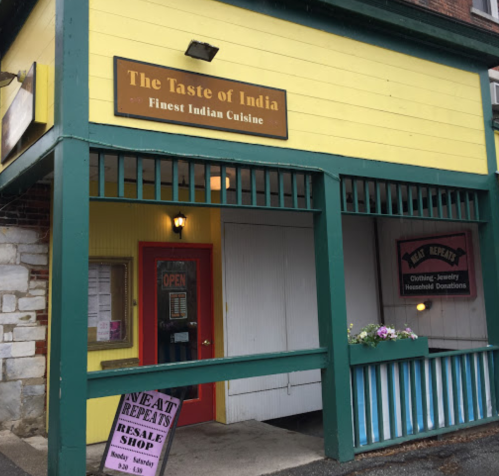 Exterior of "The Taste of India" restaurant, featuring a yellow facade and an open sign, with a resale shop nearby.