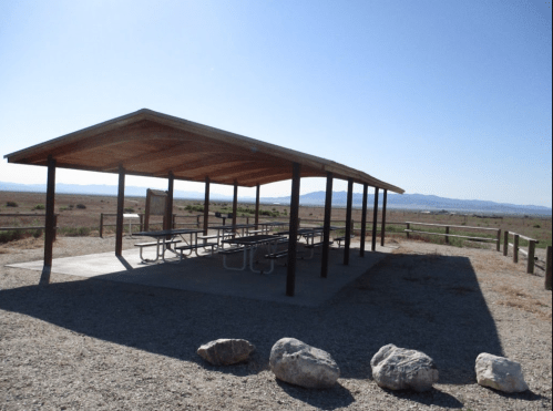 A shaded picnic shelter with tables, surrounded by a gravel area and open landscape under a clear blue sky.