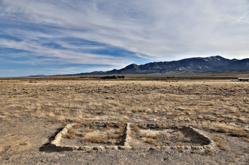 A barren landscape with two rectangular patches of dry grass, surrounded by open terrain and distant mountains under a cloudy sky.