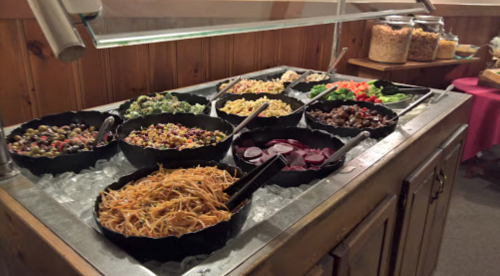 A buffet with various salads and pasta dishes in black bowls on ice, with tongs for serving.