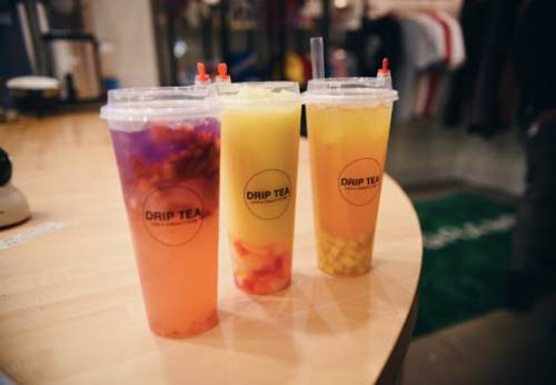 Three colorful drinks in clear cups with "DRIP TEA" labels, sitting on a wooden table in a shop.
